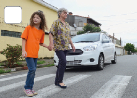 Imagem: Fotografia. Carro branco parado antes da faixa de pedestre. Na faixa há mulher idosa de cabelo curto grisalho, vestindo camiseta verde com flores roxas e calça azul. Está segurando a mão de uma menina de cabelo longo cacheado castanho, vestindo camiseta laranja e calça azul.  Fim da imagem.