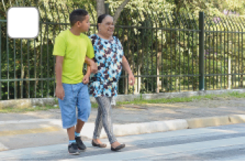 Imagem: Fotografia. Menino de cabelo curto preto, vestindo camiseta verde e bermuda azul. Está de braços cruzados com uma senhora de cabelo curto preto, vestindo camiseta azul com flores estampadas e calça preta, os dois estão andando em uma faixa de pedestre. Fim da imagem.