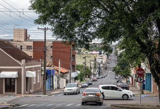 Imagem: Fotografia. Rua extensa com carros passando. Em destaque, cruzamento de ruas com placa pare e abaixo placa indicando direções, há carros cruzando a rua. Fim da imagem.