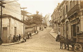 Imagem: Fotografia em tons de sépia. Rua formada por casas de dois e três andares antigas. À direita da rua, jardim alto com um muro e uma casa pequena na beirada. Sobre a rua há pessoas andando e uma charrete com caixas, puxada por dois cavalos. Fim da imagem.