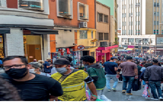 Imagem: Fotografia. Vista de rua com multidão de pessoas andando. Nas laterais há lojas abaixo de casas. Fim da imagem.