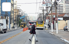 Imagem: Fotografia. Avenida dupla com duas vias. Há uma mulher de chapéu cinza, vestindo camiseta roxa e calça cinza. Em segundo plano, ônibus e carro em faixas separadas.  Fim da imagem.