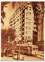 Imagem: Fotografia em preto e branco. Bondinho em uma trilha passando ao meio de uma rua com carros antigos e prédios altos. Fim da imagem.