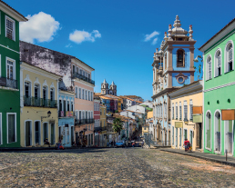 Imagem: Fotografia. Vista de ladeira de pedra com prédios baixos coloridos nos dois lados. À direita, entre os prédios e casas há uma igreja azul com duas torres laterais. Fim da imagem.