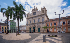 Imagem: Fotografia. Praça de pedras cinza e bege com coqueiros. Ao redor, casas coloridas e casarões grandes.   Fim da imagem.