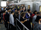 Imagem: Fotografia. Vista de multidão de pessoas em uma estação entrando em um metrô parado. Fim da imagem.