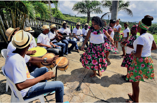 Imagem: Fotografia. Destaque de mulheres de camiseta branca e saias estampadas, dançando sobre espaço aberto. Ao redor das mulheres há homens de chapéu curto bege, camiseta branca e calça, sentados em cadeiras tocando instrumentos musicais. Fim da imagem.