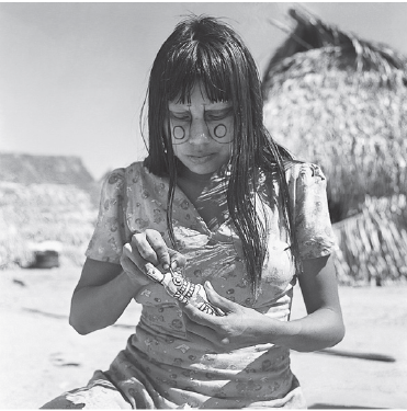 Imagem: Fotografia em preto e branco. Mulher indígena de cabelo longo e rosto com pinturas, vestindo camiseta. Atrás há uma oca de palha. Fim da imagem.