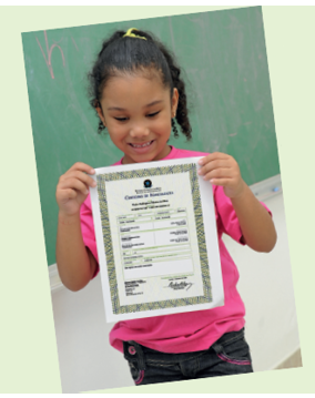 Imagem: Fotografia. Menina de cabelo longo cacheado preto, vestindo camiseta rosa. Está segurando a certidão de nascimento. Fim da imagem.