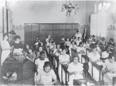 Imagem: Fotografia em preto e branco. Meninas em carteiras individuais com material sobre a mesa. Há seis fileiras de carteiras. Fim da imagem.