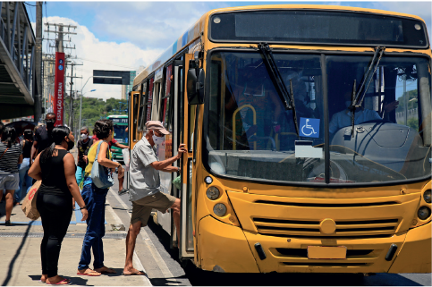 Imagem: Fotografia. Pessoas entrando em um ônibus amarelo estacionado próxima à calçada. Fim da imagem.