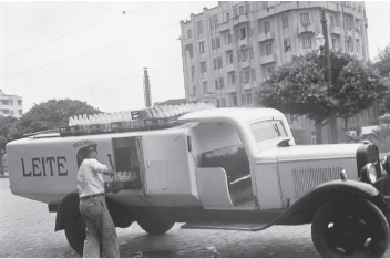 Imagem: Fotografia em preto e branco. Caminhão com caçamba fechada com portinha. Há um homem de chapéu, vestindo camiseta e calça. Está segurando uma bandeja de vidros de leite. Fim da imagem.