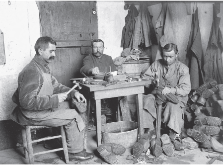 Imagem: Fotografia em preto e branco. Homens sentados ao redor de uma mesa pequena com ferramentas. Homens seguram sapatos. Ao lado, pilhas de sapatos. Fim da imagem.