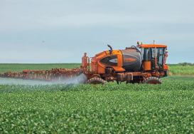 Imagem: Fotografia. Trator com tanque grande soltando líquido em jato em uma plantação extensa em campo plano. Fim da imagem.