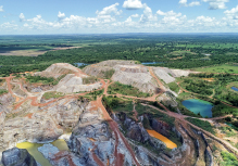 Imagem: Fotografia. Vista aérea de área de garimpo formado por morros de terra e pedra com estradas alongadas para trânsito de tratores e caminhões. Entre os morros há lagoas baixas com água suja. Ao lado, há lagoas com água azul e vegetação alta. Fim da imagem.