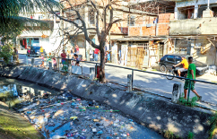 Imagem: Fotografia. Vista de córrego ao lado de rua asfaltada e casas. Sobre o córrego há lixo flutuando na água. Fim da imagem.