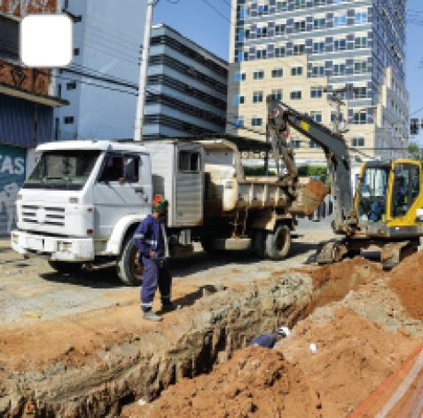 Imagem: Fotografia. Homens em obra em uma rua formando um buraco estreio e longo. Ao lado há caminhão e um trator coletando montes de terra. Fim da imagem.