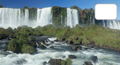 Imagem: Fotografia. Vista frontal de cachoeiras largas em queda sobre rio com pedras espalhadas e vegetação entre a água.  Fim da imagem.