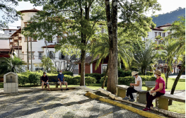 Imagem: Fotografia. Pessoas sentadas em bancos de uma praça com árvore e vegetação, ao redor há prédios baixos. Fim da imagem.