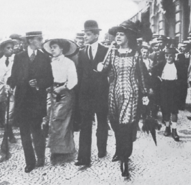 Imagem: Fotografia em preto e branco. Homens e mulheres caminhando sobre calçada. Destaque de uma mulher de chapéu de aba arredondada e cabelo longo, vestindo um vestido longo com pernas com divisões como calças. Fim da imagem.