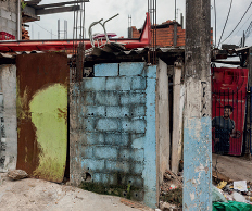 Imagem: Fotografia. Muro de tijolos a vista com ferro no lugar da porta e telha baixa sem estrutura. Fim da imagem.