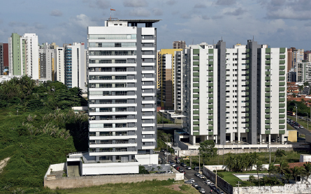 Imagem: Fotografia. Prédio com varandas cobertas por vidro de fora a fora do andar. À direita, parque com vegetação. À esquerda há prédios.   Fim da imagem.