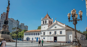 Imagem: Fotografia. Prédio grande em forma de casarão com janelas nos andares superiores. No centro há uma torre retangular com telhado triangular. À frente, pátio de concreto com monumento central. Fim da imagem.