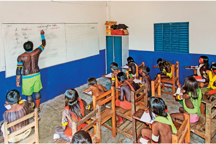 Imagem: Fotografia. Sala de aula com crianças indígenas sentadas em carteiras pequenas. À frente da lousa há um homem indígena com pinturas sobre o corpo e bermuda verde, está escrevendo sobre a lousa. Fim da imagem.