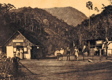 Imagem: Fotografia em preto e branco. Homens e mulheres em campo aberto com casa pequena e vegetação ao redor. Fim da imagem.