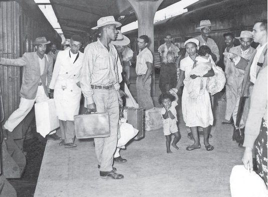 Imagem: Fotografia em preto e branco. Homens segurando malas e mulheres com crianças em uma estação de trem. Fim da imagem.