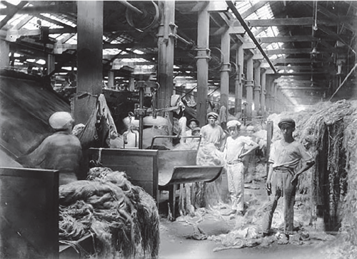 Imagem: Fotografia em preto e branco. Homens e mulheres trabalhando interior de uma fábrica com maquinários altos com restos de tecidos e lã. Fim da imagem.