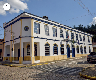 Imagem: Fotografia. 1: Prédio branco de esquina com detalhe em amarelo, portas e janelas azuis. Ao lado, rua de pedra com faixa de pedestres. Fim da imagem.