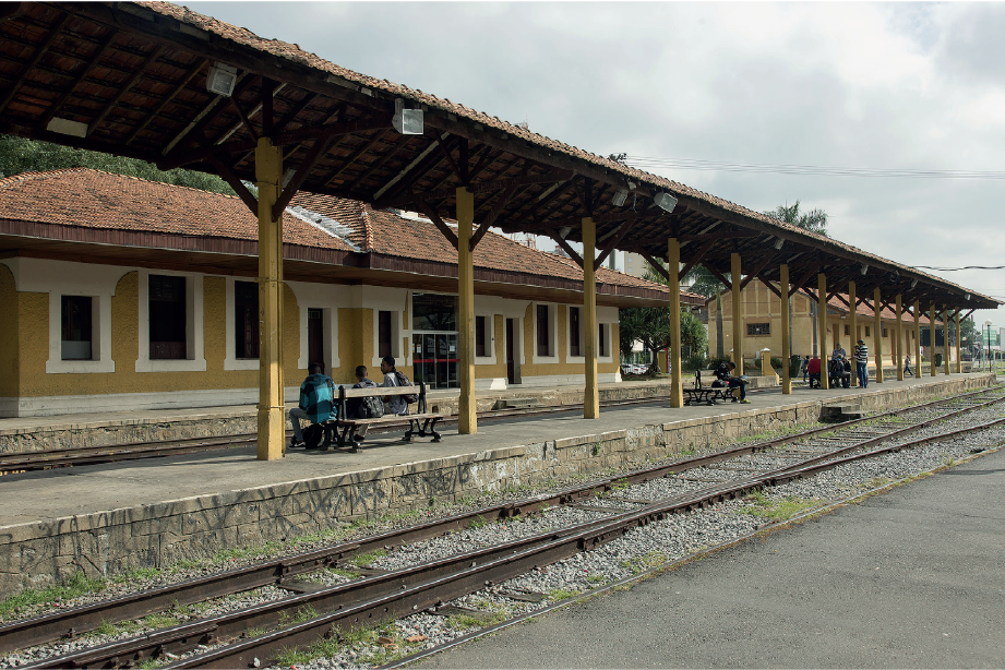 Imagem: Fotografia. Vista de estação ferroviária com trilho ao lado de passarela. Na passarela há vigas de madeira sustentando um telhado triangular. Atrás há casa de parede amarela e branca. Entre as vigas há bancos. Fim da imagem.