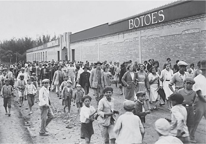 Imagem: Fotografia em preto e branca. Vista de multidão de pessoas andando, predominância de crianças na fotografia.  Ao lado há um barracão indicando no letreiro: “botões”. Fim da imagem.