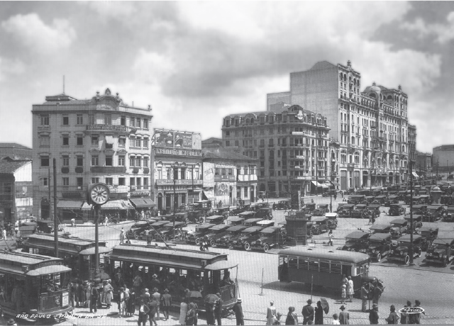 Imagem: Fotografia em preto e branco. Vista de rua com prédios altos e carros antigos em estacionamento na lateral. Há ônibus passando nas ruas e aglomerado de pessoas entre veículos e ônibus. Fim da imagem.