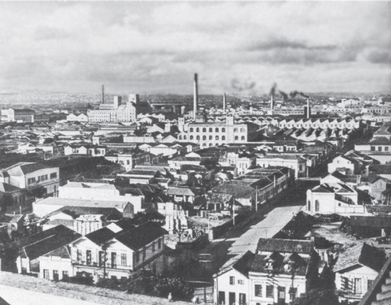 Imagem: Fotografia em preto e branco. Vista aérea de cidade plana com casas, poucas árvores e ao fundo indústrias soltando fumaça por chaminés altas. Fim da imagem.