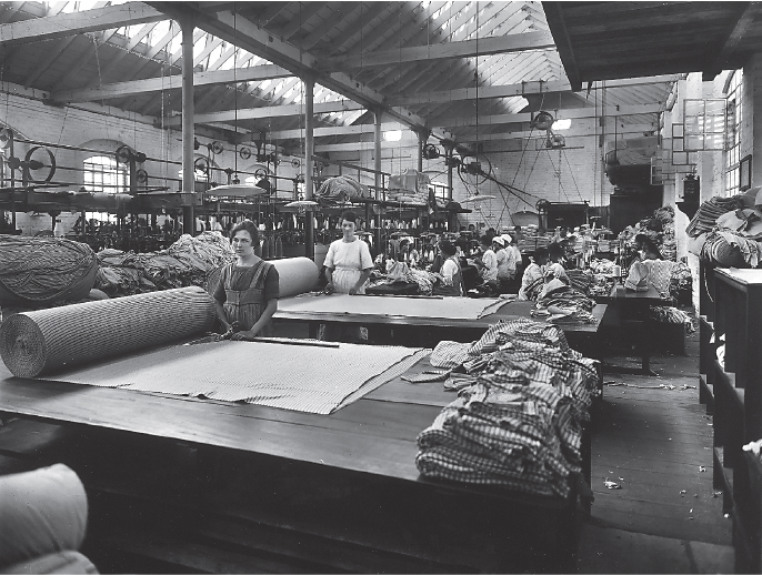 Imagem: Fotografia em preto e branco. Linha de produção com mesas longas com pilhas e rolos de tecidos. Há mulheres atrás de cada mesa. Ao fundo do galpão, mulheres sentadas em frente a máquinas de costura. Fim da imagem.