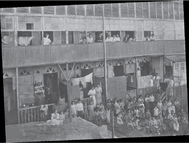 Imagem: Fotografia em preto e branco. Vista de cortiço de dois andares. Há multidão de pessoas em varanda e área central na frente. Há pequenas portas lado a lado com muro baixo dividindo os apartamentos. Fim da imagem.