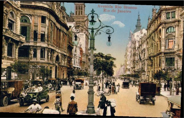 Imagem: Fotografia. Vista de avenida com duas vistas separadas por postes antigos com lâmpadas largas em três pontos. Em ambos os lados há prédios altos de estrutura antiga. Fim da imagem.