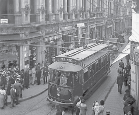 Imagem: Fotografia em preto e branco. Bonde passando em rua estreita com trilho central e antena ligada a um cabo superior que acompanha o trilho. Ao redor da rua há pessoas em pé. Fim da imagem.