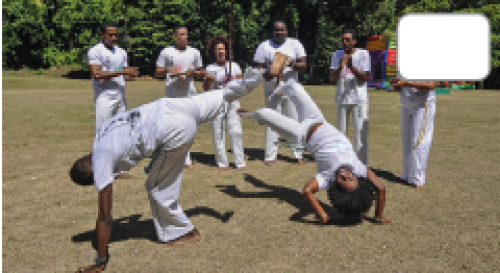 Imagem: Fotografia. Homens e mulheres em um campo, todos vestindo roupas brancas, jogando capoeira. Há homens no centro em movimento e homens ao redor tocando instrumentos musicais. Fim da imagem.