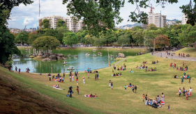 Imagem: Fotografia. Vista de parque com gramado e lago central. No gramado há grupos de pessoas sentadas. Fim da imagem.