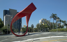 Imagem: Fotografia. Destaque de uma escultura no centro de uma praça. A escultura possui um círculo irregular curvado vermelho.  Fim da imagem.