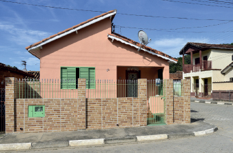 Imagem: Fotografia. Vista de casa de esquina de madeira rosa com janela verde, há muros de tijolos e grade baixa. Fim da imagem.