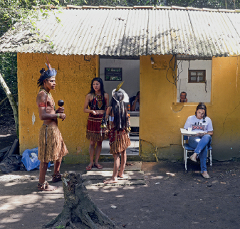 Imagem: Fotografia. Homem indígena de cabelo curto preto, vestindo tanga de palha e cocar colorido, e duas mulheres indígenas de cabelos longos preto, vestindo vestido de palha colorida e cocar colorido, os três estão em frente a uma casa amarela de alvenaria. Ao lado da porta, mulher de cabelo longo preto, vestindo camiseta branca e calça azul, sentada em uma carteira individual. Fim da imagem.