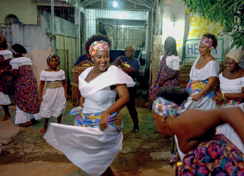 Imagem: Fotografia. Mulheres de vestido branco com pequenas estampas, dançando em círculos na rua. Fim da imagem.