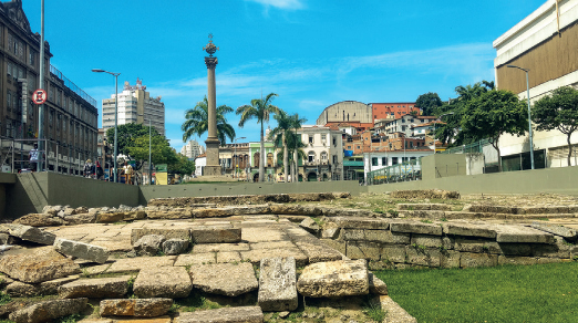 Imagem: Fotografia. Vista de pedras largas formando estrutura baixa de construção antiga. Ao redor há casas e pessoas circulando. Fim da imagem.