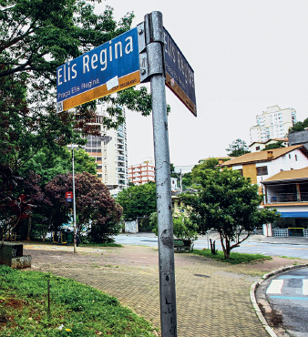 Imagem: Fotografia. Destaque de placa em uma praça indicando “Elis Regina”. Na praça há arvores, bancos e calçada entre vegetação. Fim da imagem.