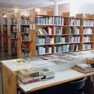 Imagem: Fotografia. Vista de uma biblioteca com prateleiras longas de madeira com livros. Fim da imagem.