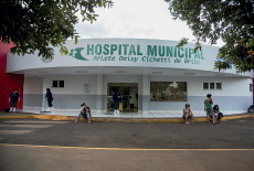 Imagem: Fotografia B: Prédio branco com pessoas sentadas na calçada e no seu interior. Há uma placa indicando “hospital municipal – Arlete Daisy Cichetti de Brito” Fim da imagem.